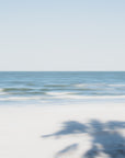 Beach scene with palm tree shadow on sand and clear blue sky.