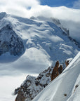 A panoramic view of snow-covered mountains, with Mont Blanc prominently featured, cloudy skies above, and a rocky outcropping on the side.