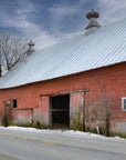 A red barn with a gray roof, situated on a roadside with a winter landscape in the background, including trees and a silhouette of mountains.