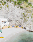 A scenic view of a cove with clear blue water and a rocky shoreline. In the background, there is a small seaside town with colorful boats docked on a sandy beach, and a white house on the Italian coast
