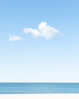 Minimal beach scene with blue sky and one cloud in the sky over a calm ocean.